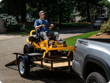 Man with hat and blue shirt driving a yellow lawn mower onto a trailer