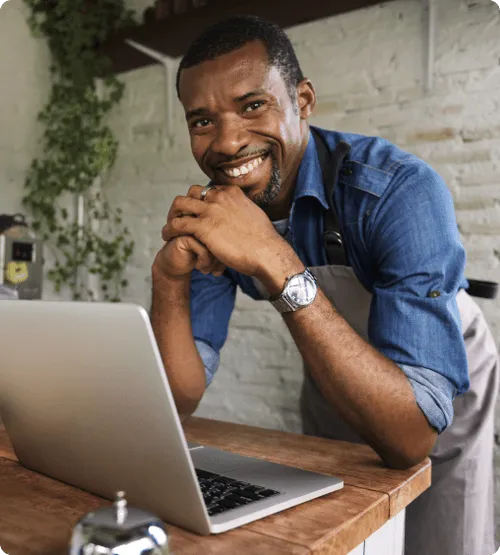 A smiling man in a blue shirt and apron leaning over a table with a laptop