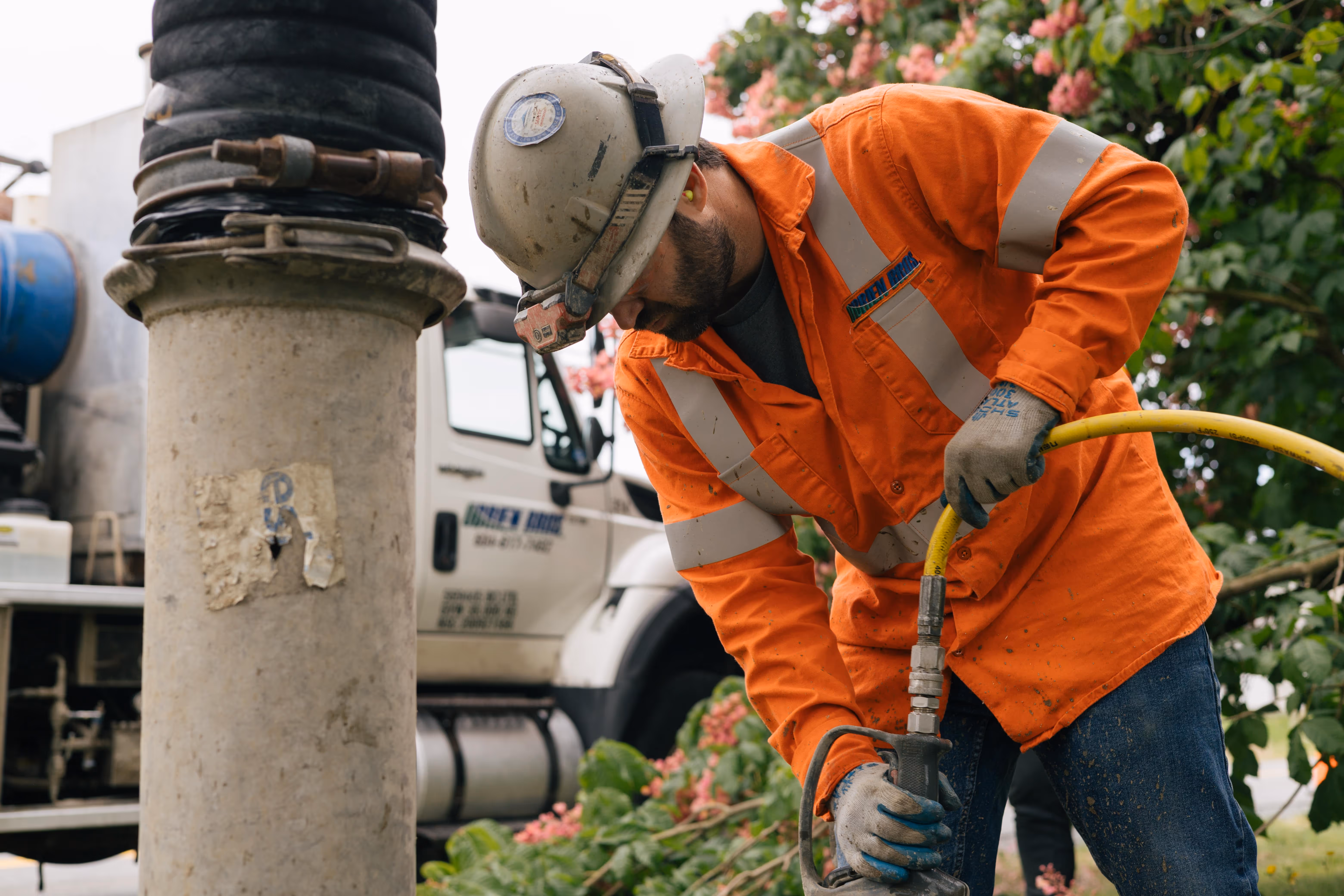 Construction worker in orange safety gear operates heavy equipment outdoors near a truck.