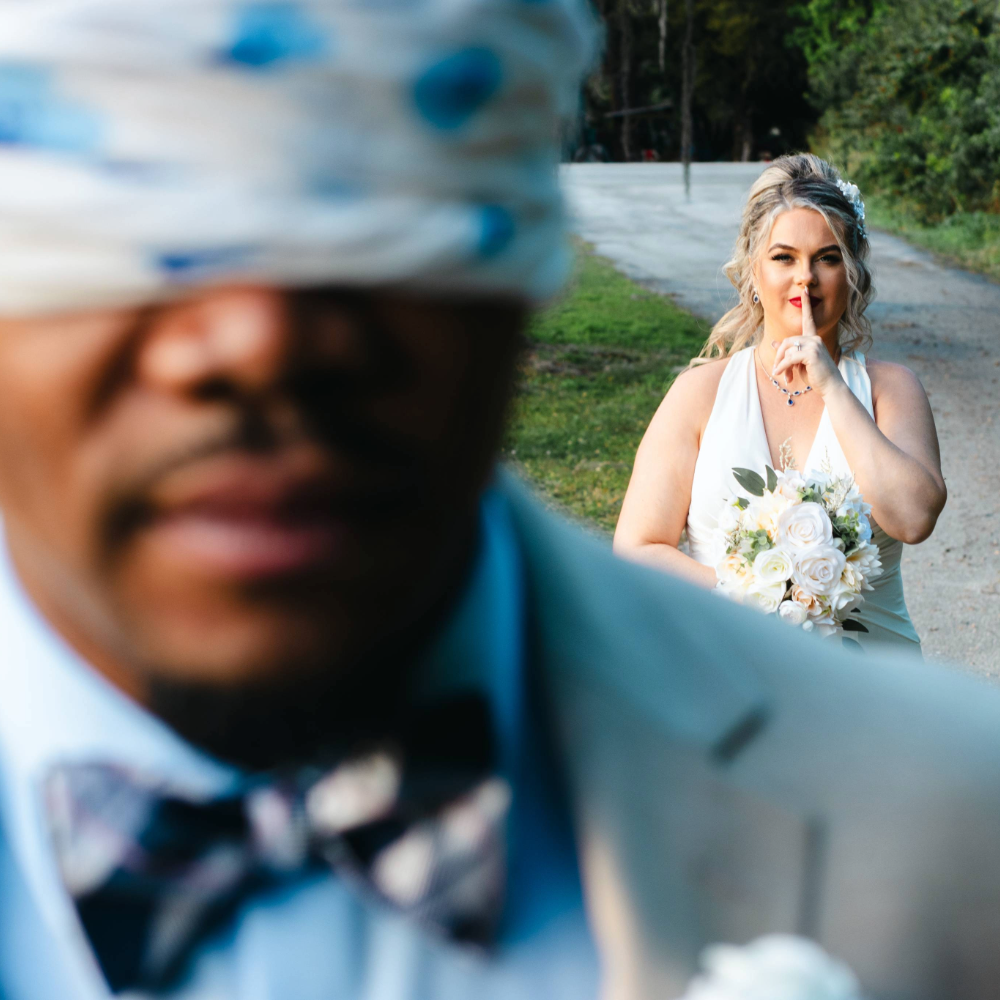 A groom being blindfolded while the bride is standing behind him holding a bouquet. 
