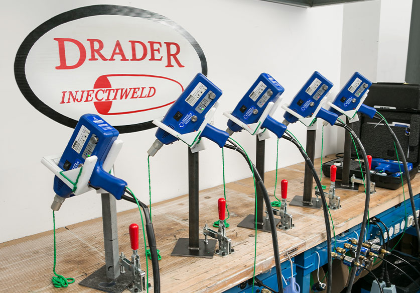 Row of five blue Drader Injectiweld devices mounted on stands along a wooden workbench with green cords and red handles.