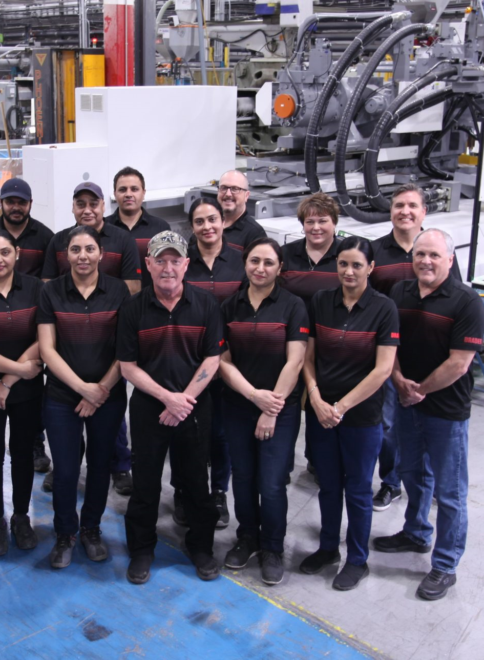 Group of factory workers wearing black and red company shirts standing in an industrial workspace with machinery in the background.