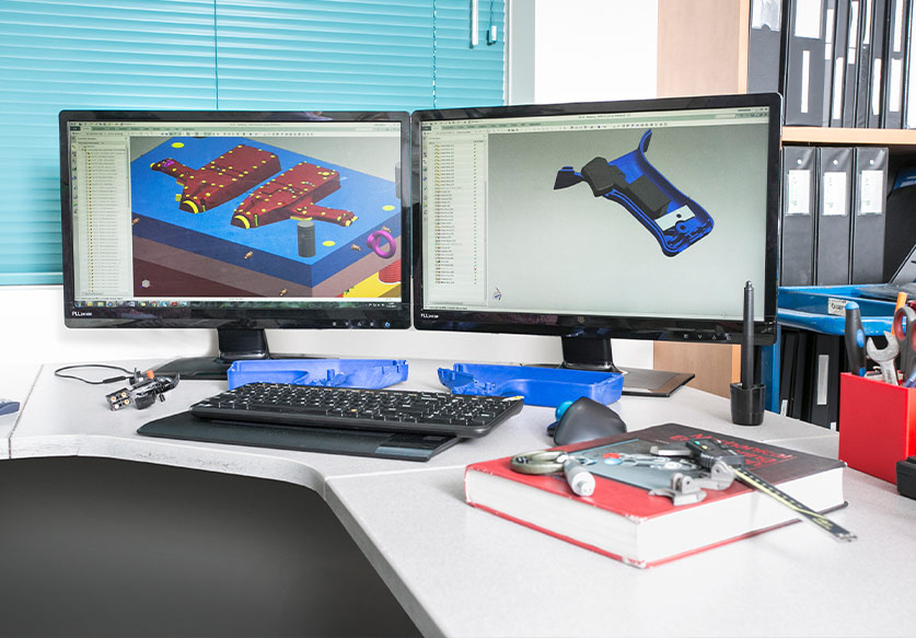 Dual computer monitors on a desk displaying 3D CAD designs with a keyboard, mouse, and engineering tools on the desk surface.