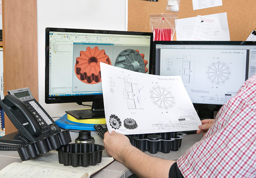 Person reviewing technical drawings of a gear-like component with corresponding 3D models displayed on two computer monitors.