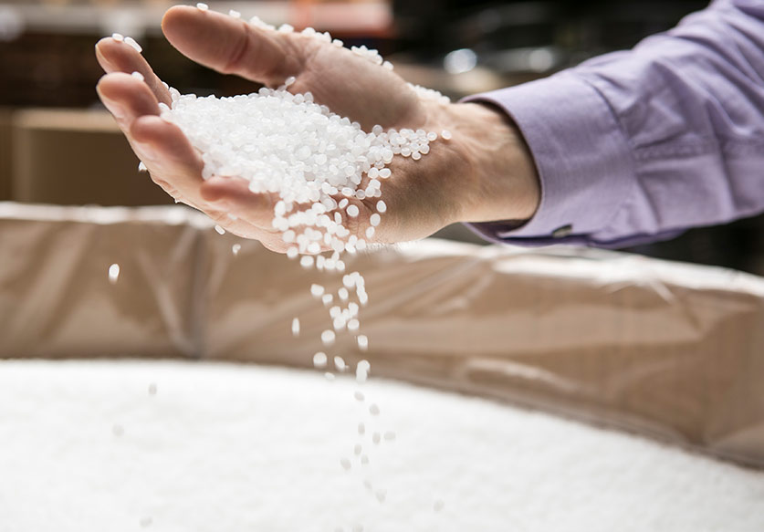 Hand holding and letting small white plastic pellets fall over a large container filled with the same pellets.