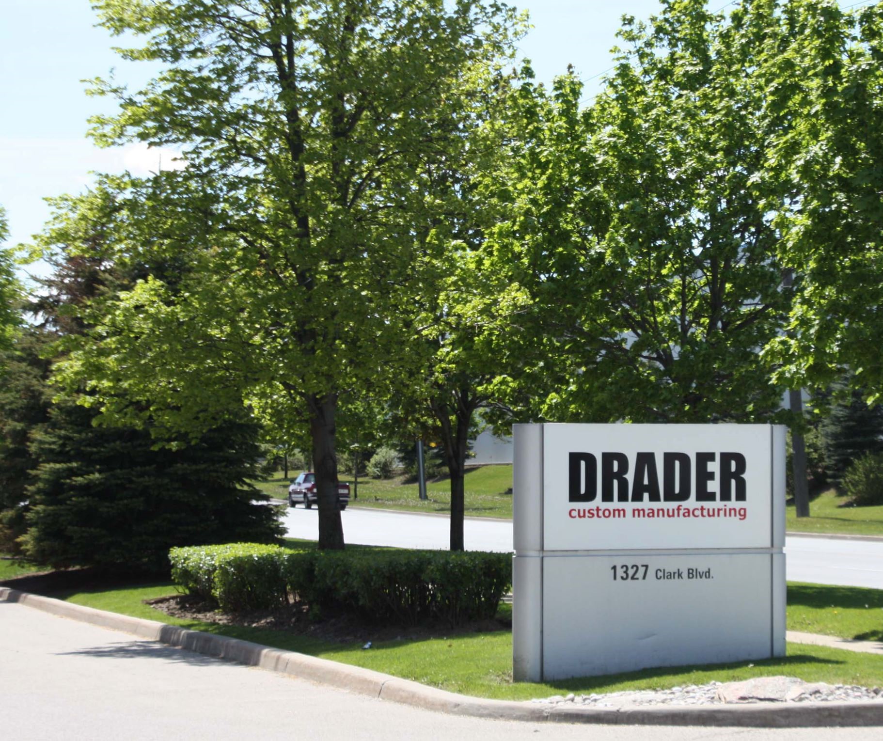 Roadside sign for Drader custom manufacturing at 1327 Clark Blvd, surrounded by green trees and shrubs.