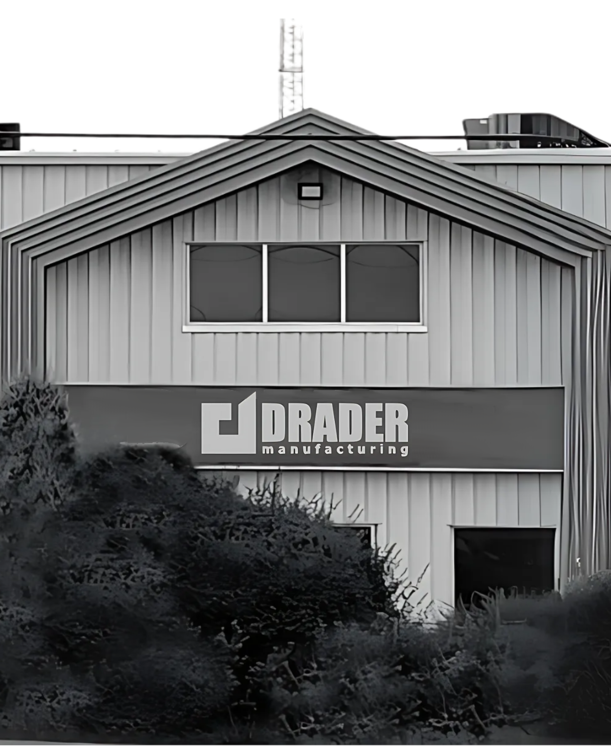 Metal warehouse building with a DRADER manufacturing sign on the front and bushes in the foreground.