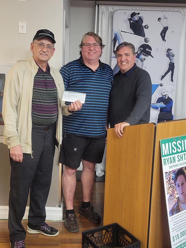 Three men standing indoors; the man in the middle holds a check, and the man on the right leans on a podium with a missing person poster.