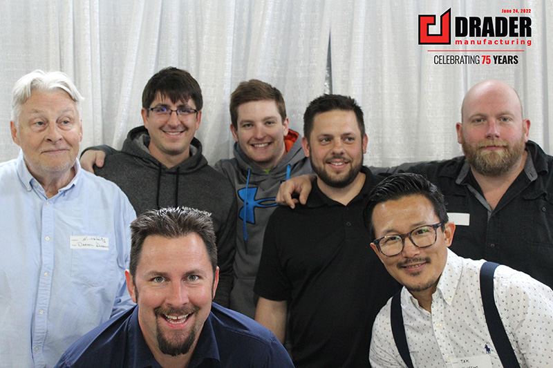 Group of seven men smiling in front of a white curtain with Drader Manufacturing logo celebrating 75 years.