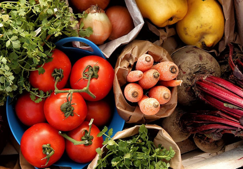 Fresh vegetables including tomatoes in a blue basket, carrots in a paper bag, onions, yellow pears, beets with red stems, and leafy greens.