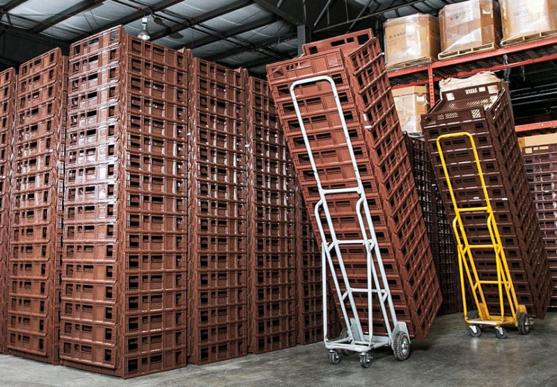 Stacked brown plastic storage crates in a warehouse with two hand trucks, one gray and one yellow, leaning against the crates.