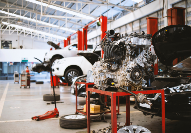 Car engine mounted on a red metal stand in a busy automotive repair shop with cars on lifts and tires on the floor.