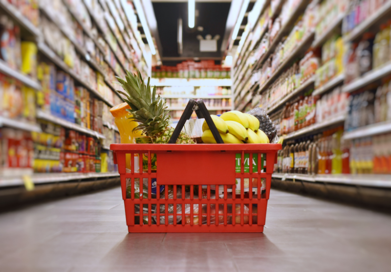 Red shopping basket filled with fruits including a pineapple, bananas, and beverages in a grocery store aisle.