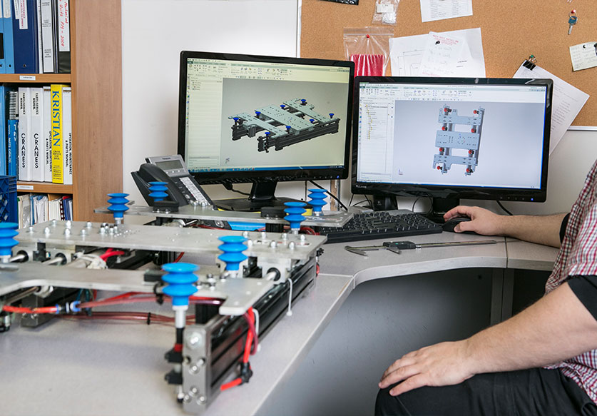 Engineer working at a desk with dual monitors displaying 3D CAD designs of a mechanical assembly, with a physical prototype on the desk.