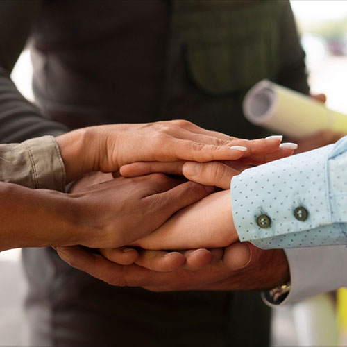 Close-up of multiple hands stacked together in a gesture of unity, with people in business and casual attire in the background.
