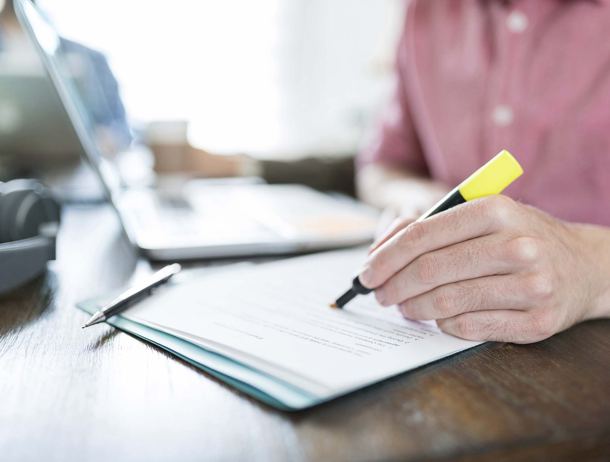 A person writing on a document with a yellow highlighter at a wooden desk, with a laptop, pen, and headphones in the background.