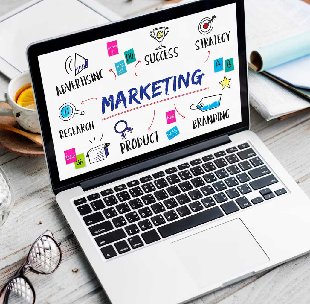 A laptop displaying a colorful marketing diagram with terms like "Advertising," "Research," "Product," "Branding," "Strategy," and "Success," placed on a wooden table with a cup of tea, glasses, and notebooks nearby