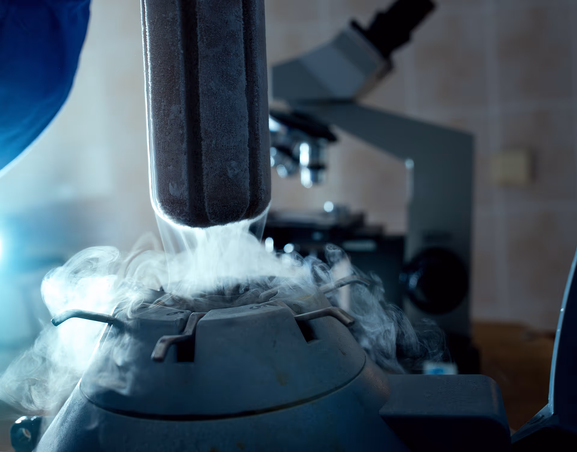Close-up of a cryogenic container releasing vapor with a microscope in the background.