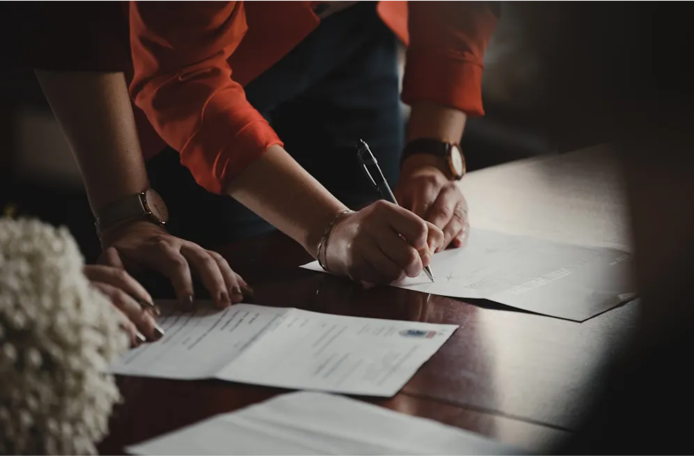 A person signing documents on a wooden table, highlighting the importance of paperwork in business transactions.