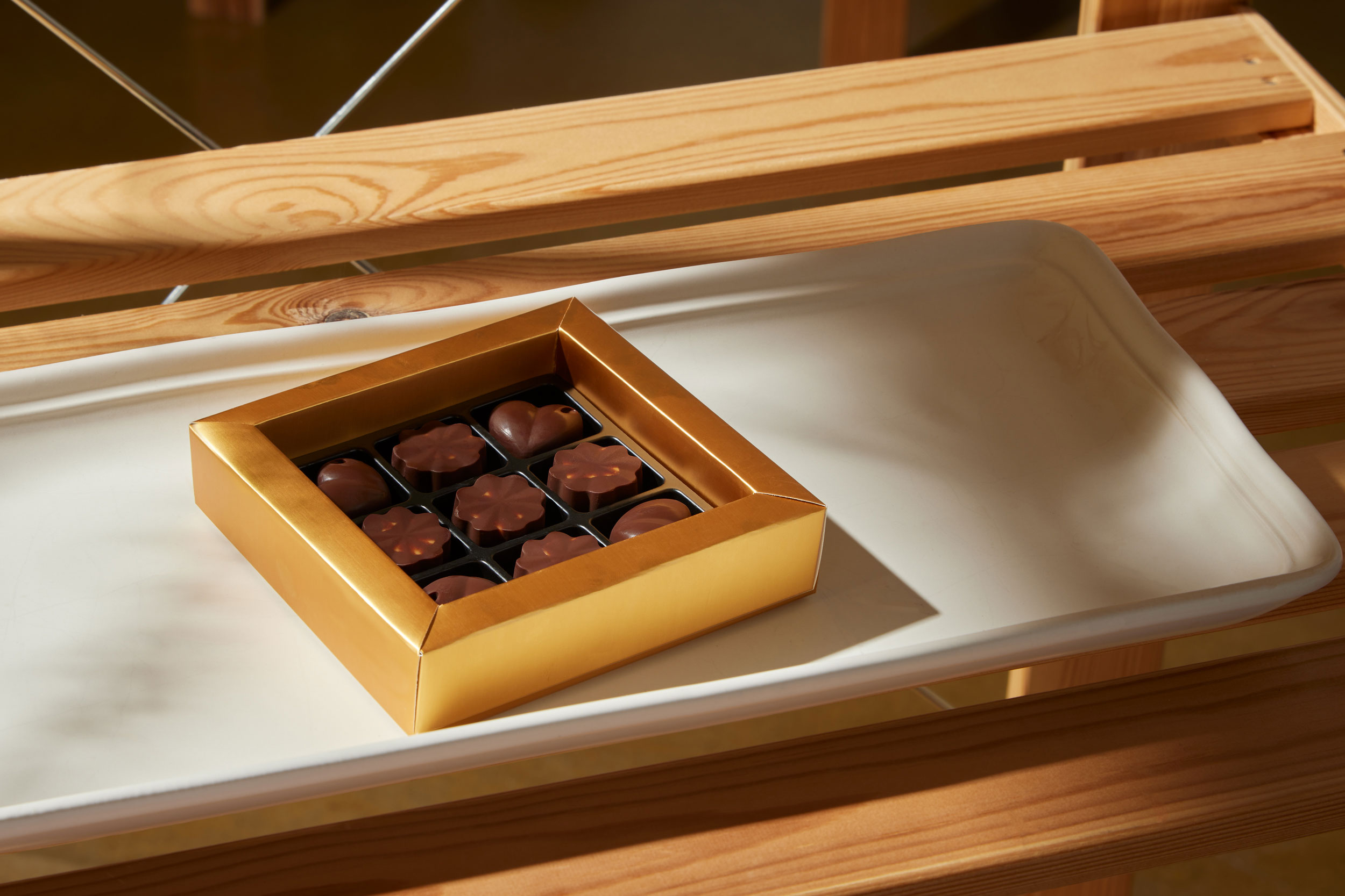 A gold gift box of assorted chocolates, including heart and flower-shaped pieces, displayed on a white ceramic tray with a wooden background.