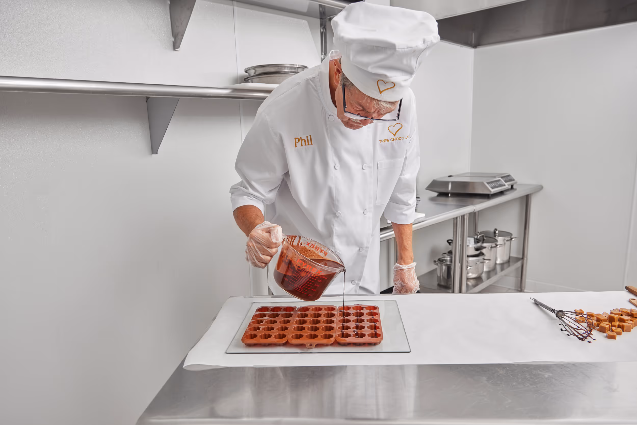 A chocolatier named Phil, wearing a white chef’s uniform and hat with the Trew Chocolate logo, pours liquid chocolate into silicone molds in a professional kitchen.