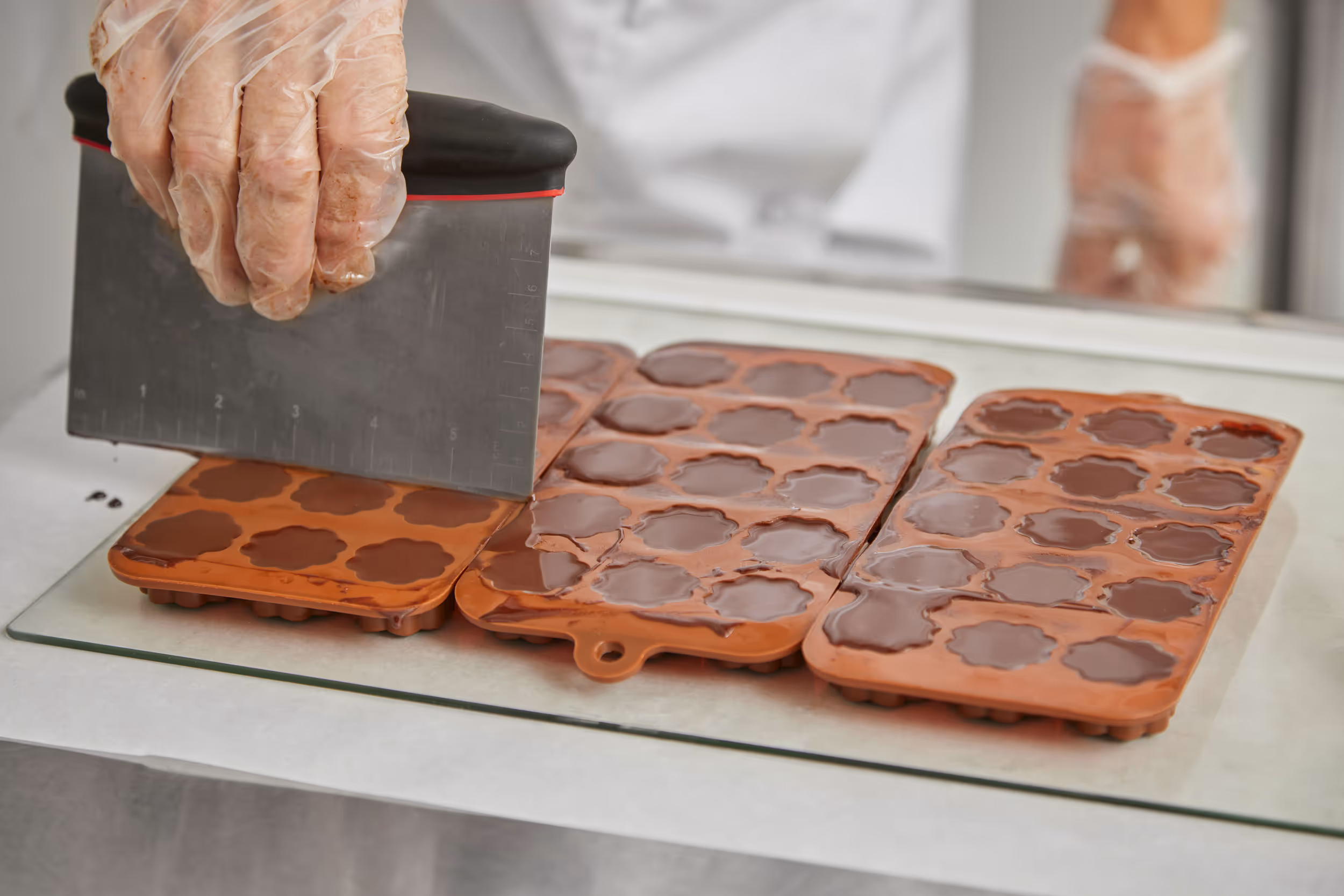 Close-up of a chocolatier smoothing liquid chocolate in silicone flower-shaped molds using a metal scraper in a professional kitchen.