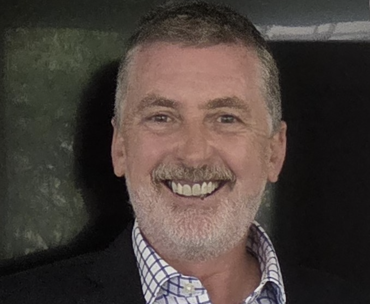 Smiling middle-aged man with short gray hair and beard wearing a checked dress shirt and dark jacket.