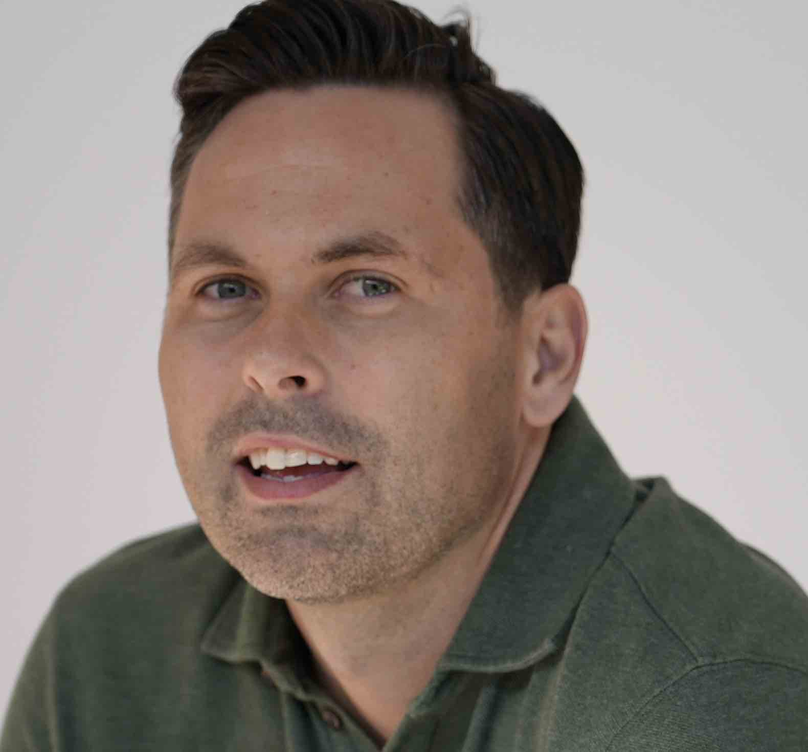 Close-up of a man with short dark hair and light stubble wearing a green collared shirt, looking slightly to the side.