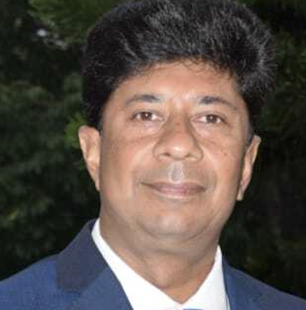 Man wearing a dark blue suit jacket and white shirt, posing outdoors with greenery in the background.
