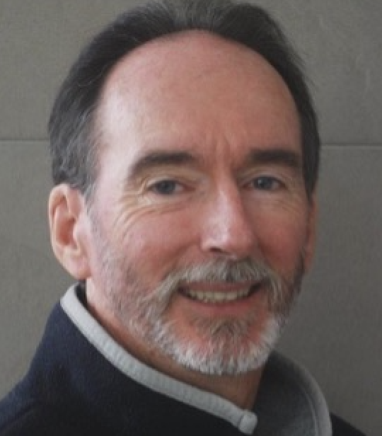 Middle-aged man with gray hair and beard smiling, wearing a dark jacket with gray collar.