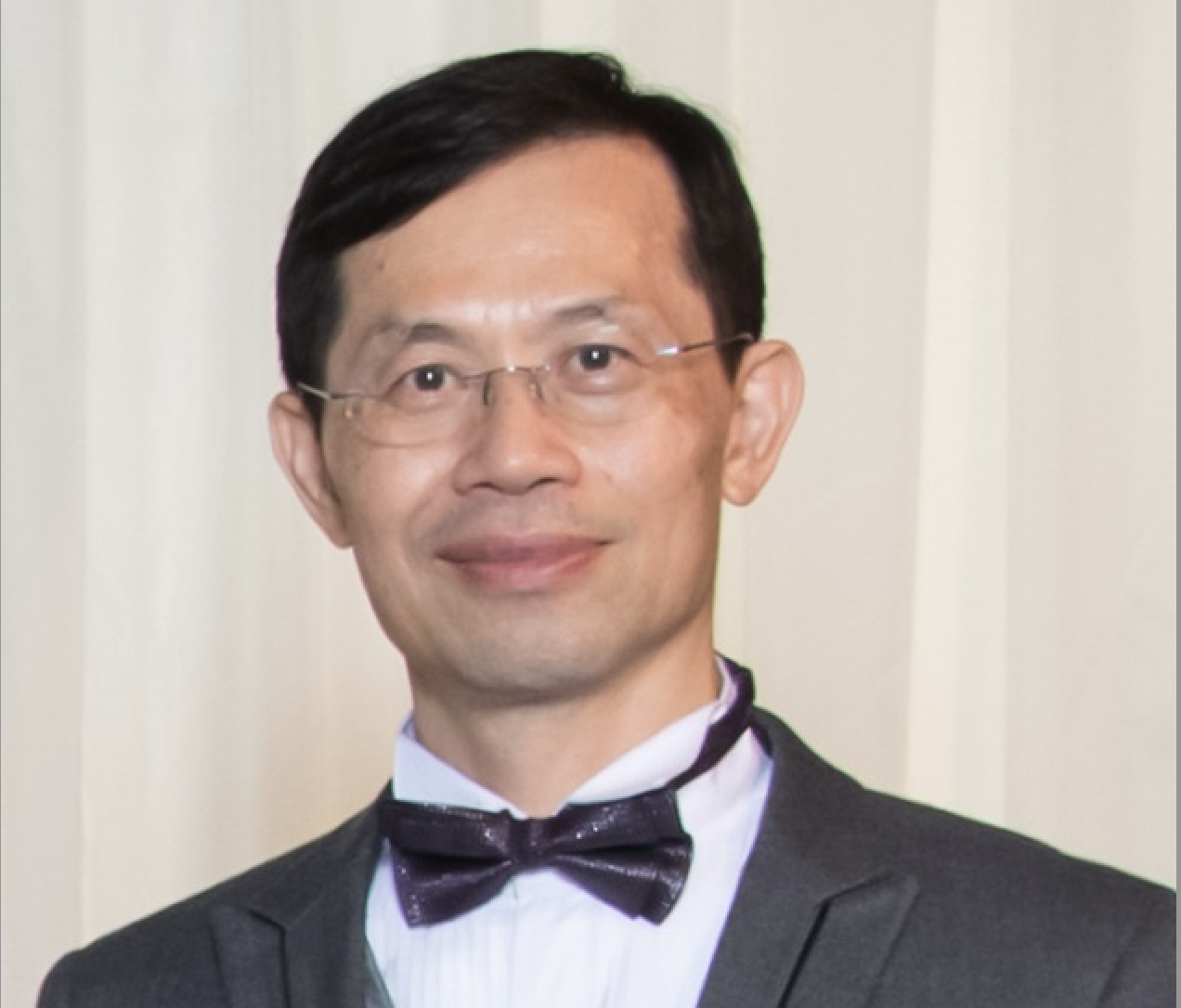 Man wearing glasses, a dark bow tie, white shirt, and dark suit jacket in front of a light background.