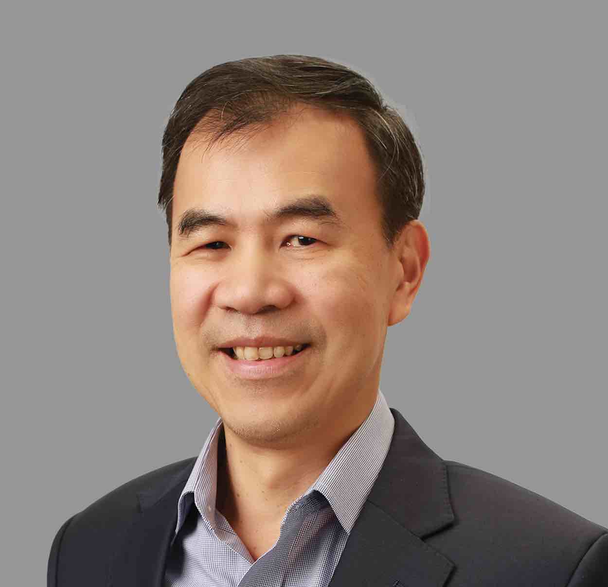 Smiling middle-aged Asian man wearing a dark blazer and light blue collared shirt against a gray background.