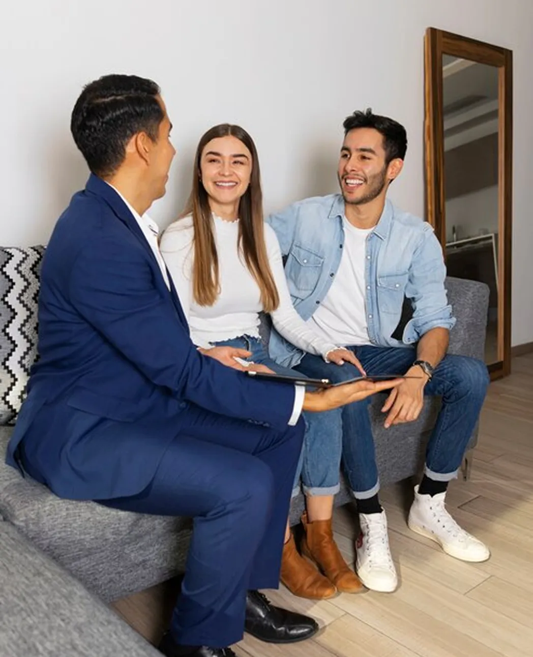 Mortgage broker in navy suit discussing with smiling young couple seated on grey couch.