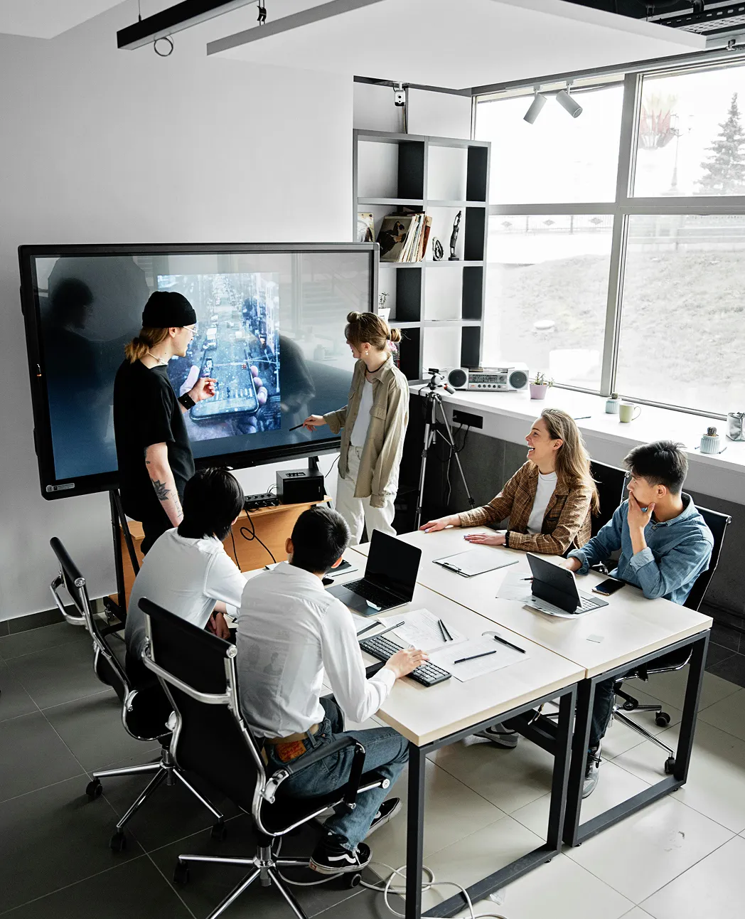 Five people in a meeting room watching a large screen showing a smartphone with a city view.