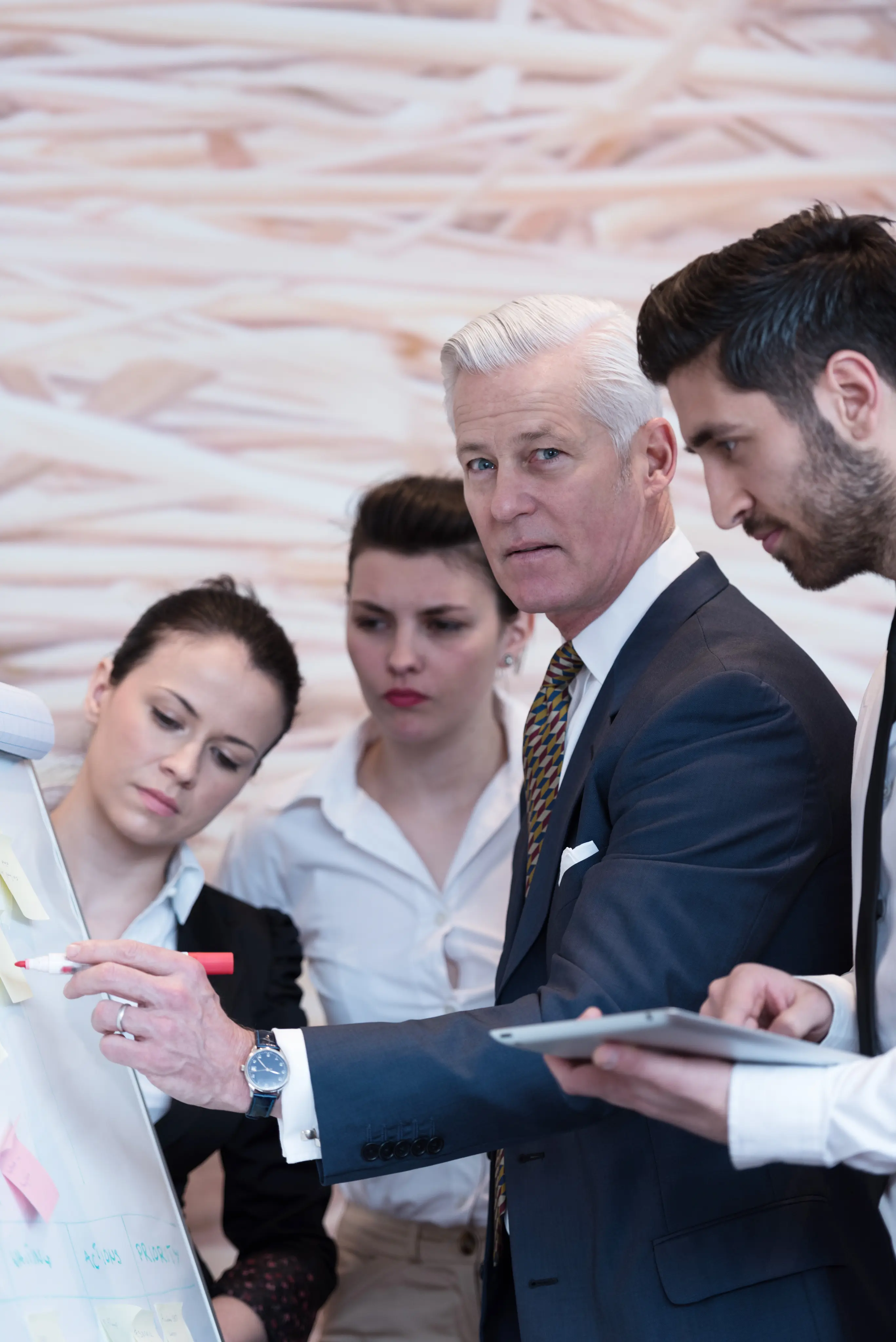 A mature man in a navy suit points with a red marker at a flip chart covered with sticky notes while three colleagues attentively observe, including a woman in a black jacket, a woman in a white shirt, and a man holding a tablet.
