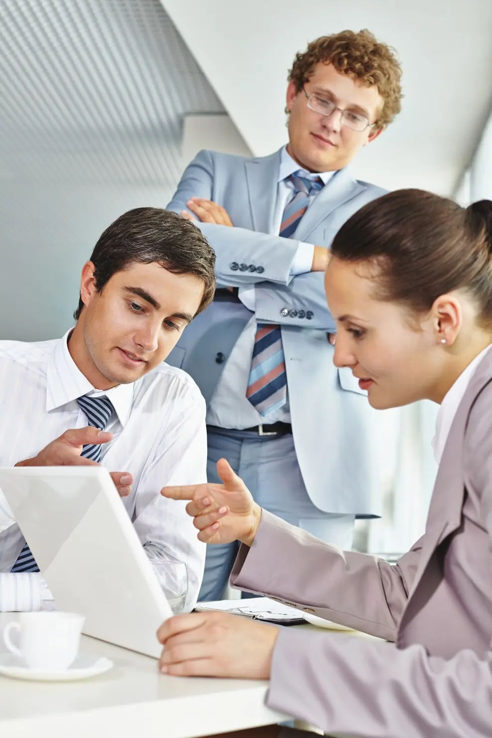 Three business professionals in an office setting are looking at a tablet device; a woman in a light blazer points at the screen while a man in a white shirt and striped tie views it attentively; a third man in a light blue suit and glasses stands behind them with arms crossed, observing.