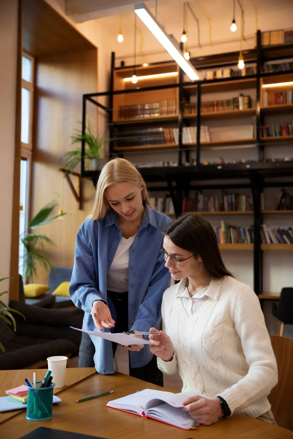 Two women in a library or study room working together while reviewing papers; one woman with blonde hair stands pointing at the documents, and the other with dark hair and glasses sits holding a pen and notebook.