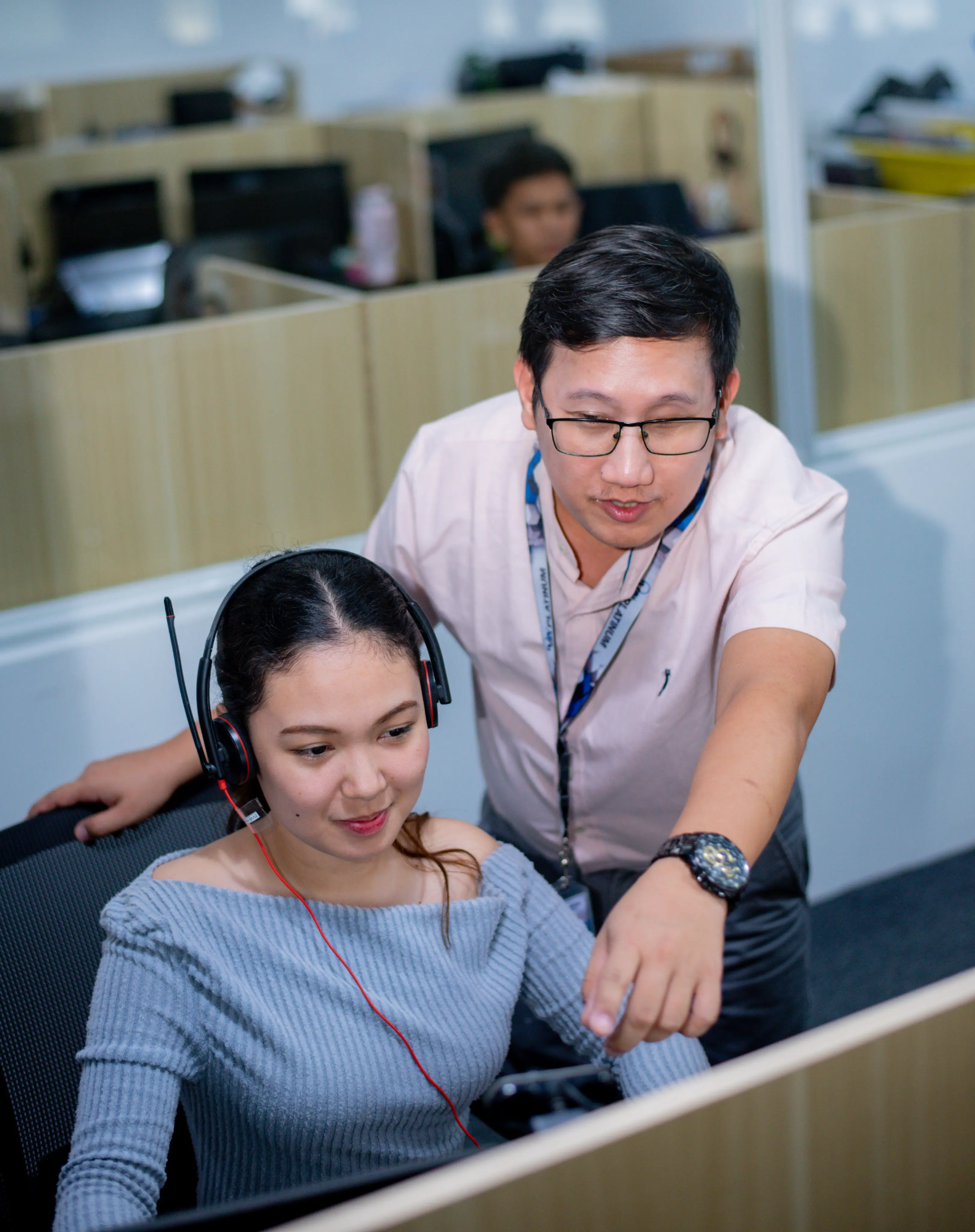 A man wearing glasses and a lanyard points at a computer screen while standing next to a woman wearing a headset and a blue off-shoulder top, seated in an office cubicle.