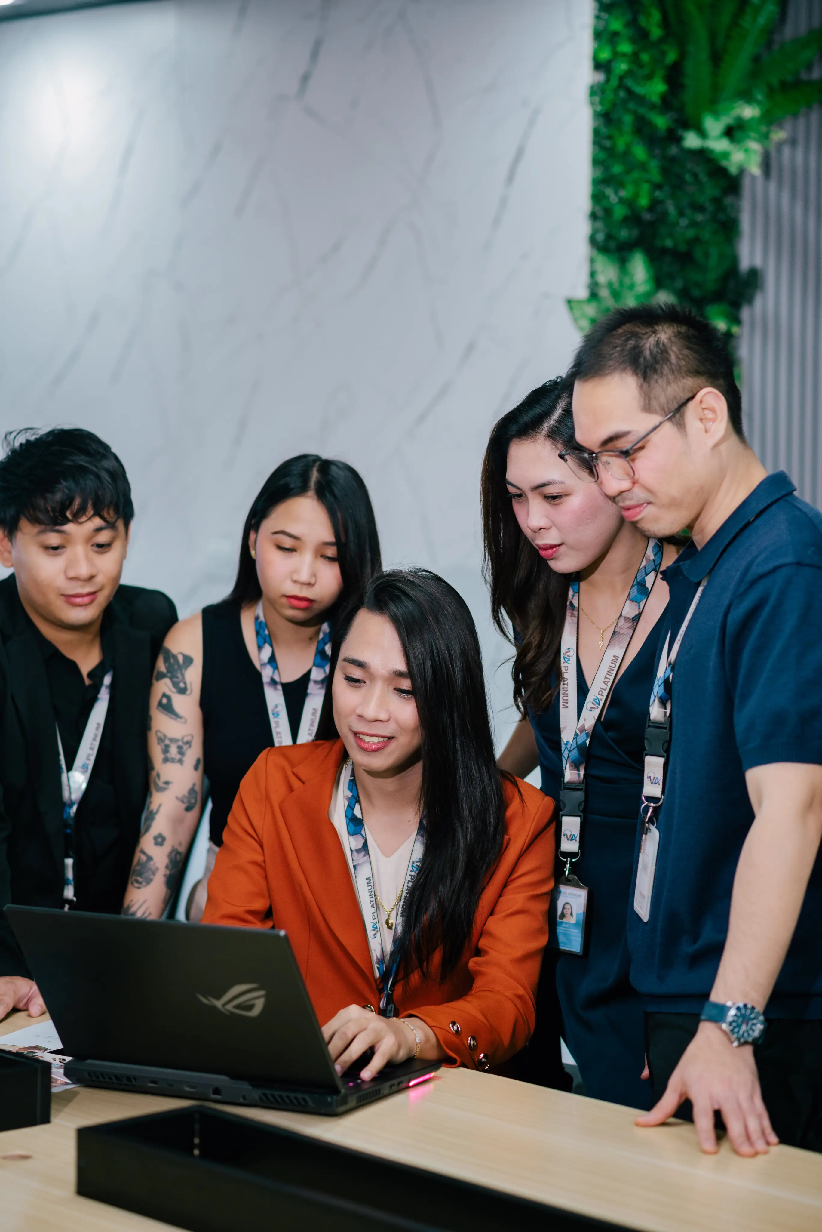 Five colleagues gathered around a laptop on a table in an office setting; one person in an orange blazer is using the laptop while others watch attentively.