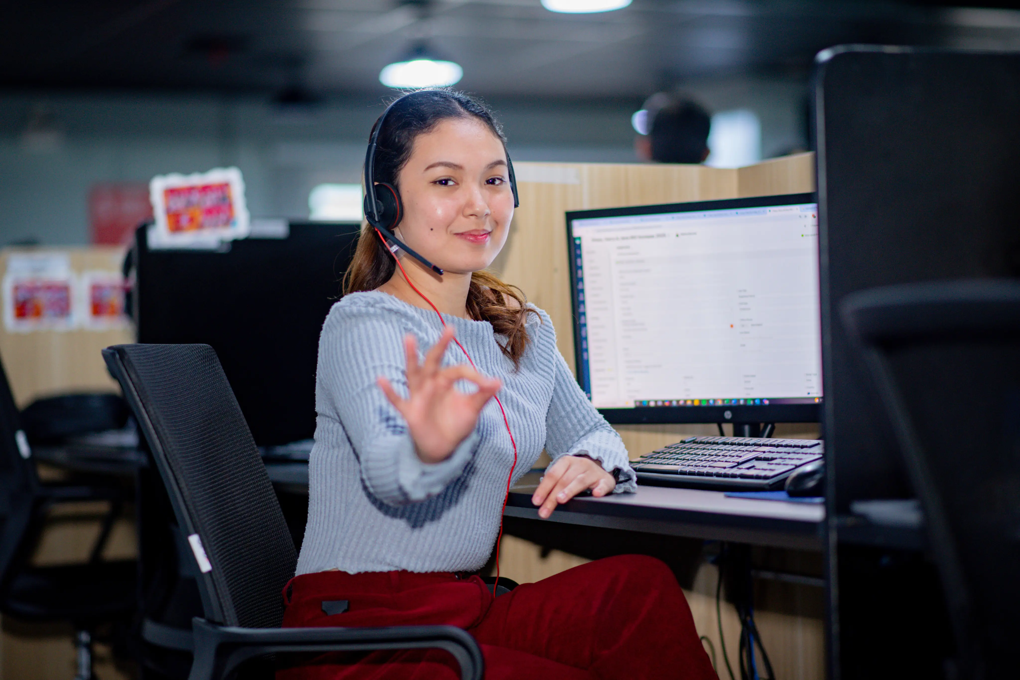 A woman wearing a headset sits at a desk in an office cubicle, smiling and making an OK gesture with her hand, with a computer monitor and keyboard in front of her.