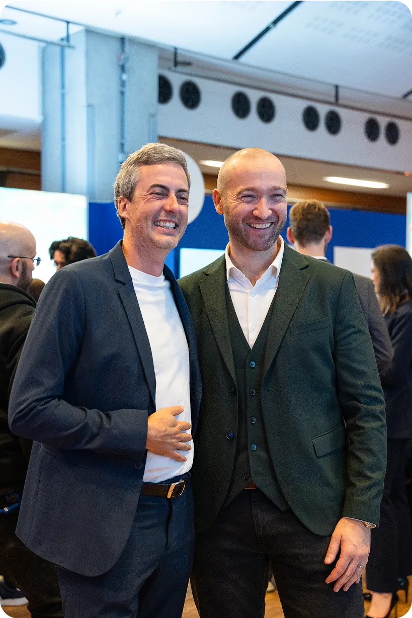 Nicolas Chéron posant avec un autre homme en costume lors d’un événement professionnel, tous deux souriants devant un fond animé.