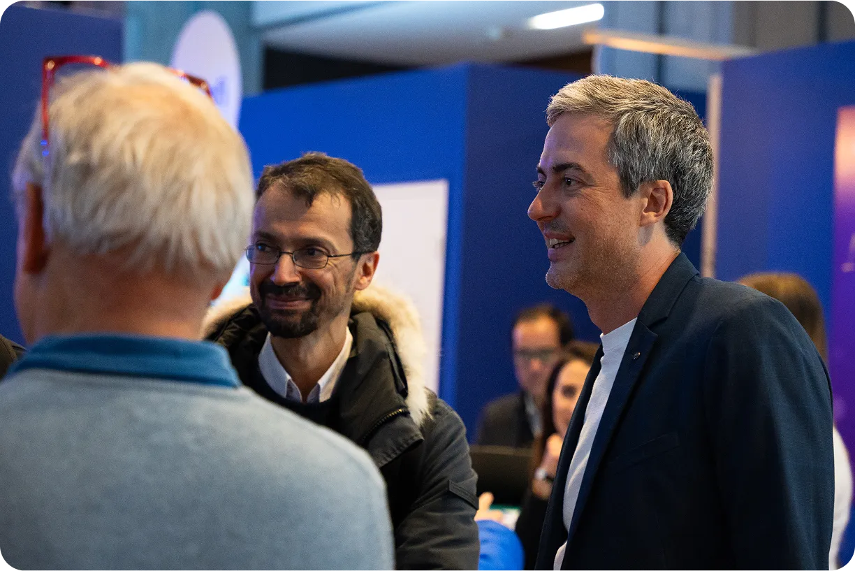 Nicolas Chéron souriant, en pleine conversation avec deux autres hommes dans un espace d’exposition au fond bleu.
