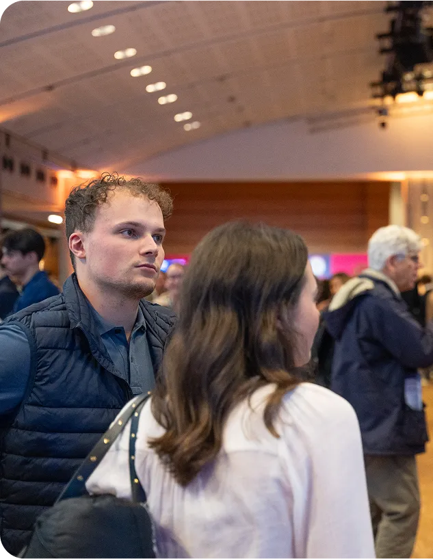 Jeune homme en doudoune bleue échangeant avec une femme dans un espace événementiel lumineux, entourés d’autres participants.