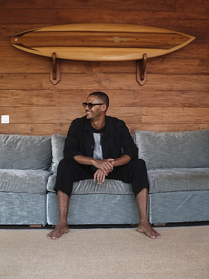 Simo Azzaoui sitzt barfuß auf einem grauen Sofa vor einer hölzernen Wand mit einem an der Wand montierten Surfbrett.