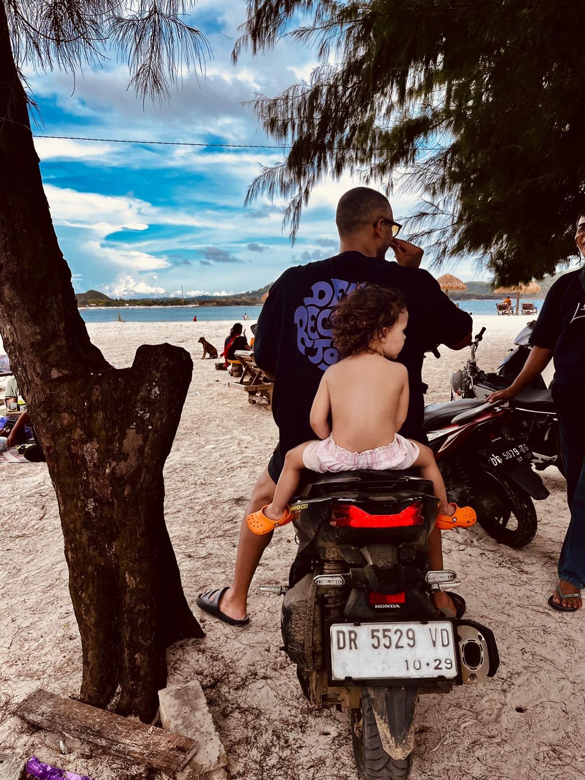 Simo-Azzaoui sitzt mit seiner Tochter auf einem Motorrad am Strand mit Blick auf Meer und bewölkten Himmel.