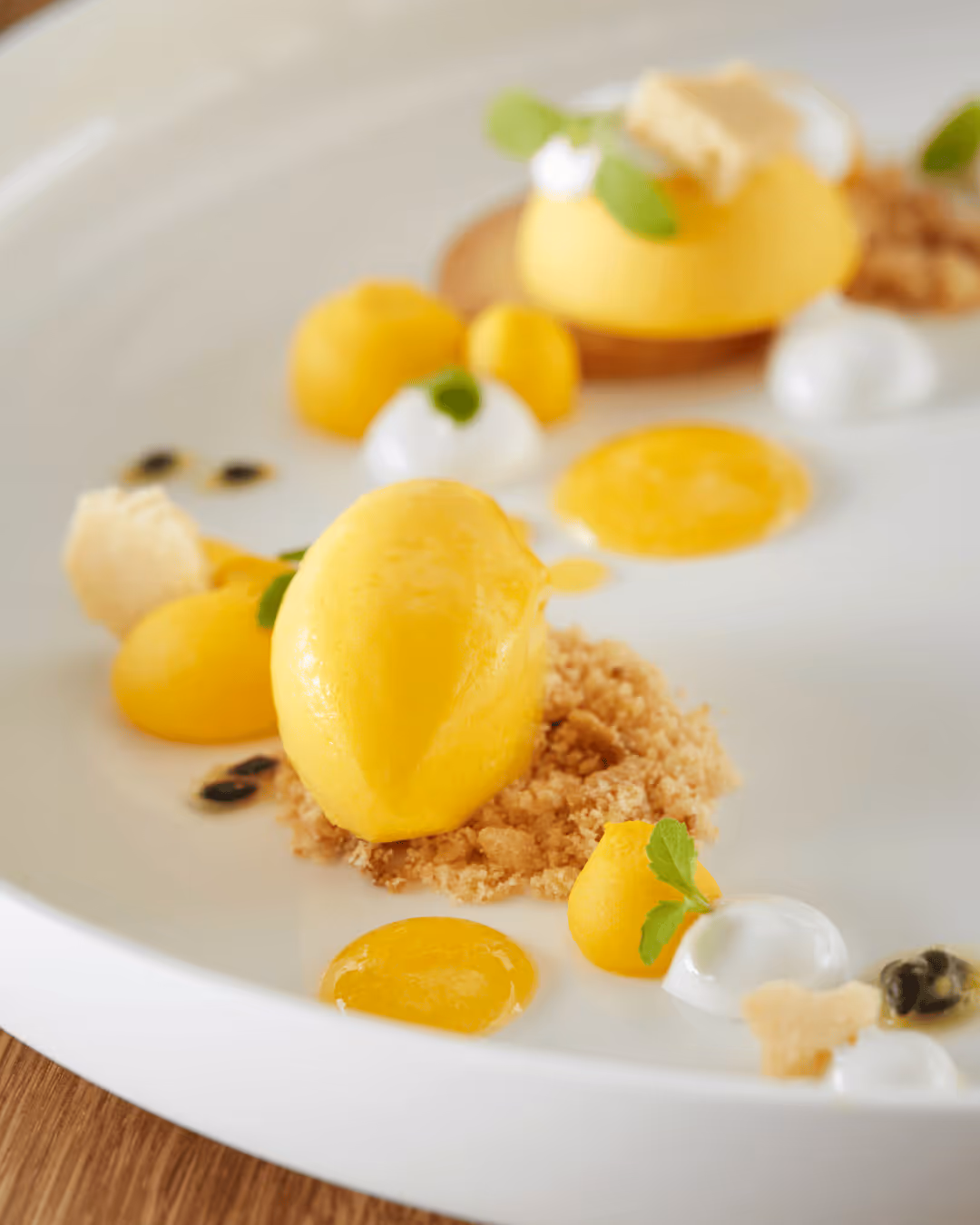 Artfully prepared dessert with yellow sorbet, crumble, fruit cream and fine garnish on a white plate.