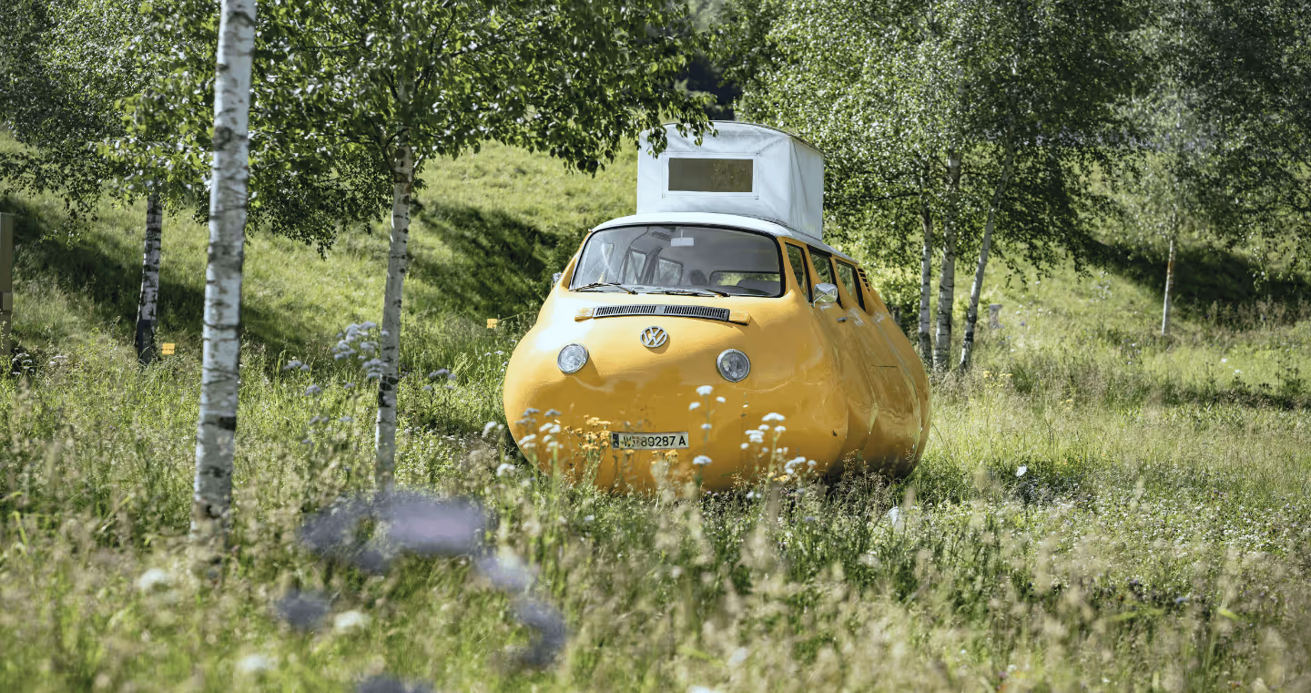 A bright yellow, rounded VW bus with a white pop-up roof stands in the park of Swarovski Kristallwelten, surrounded by trees. The playful vehicle is an art installation by Erwin Wurm and fits humorously into the natural setting.