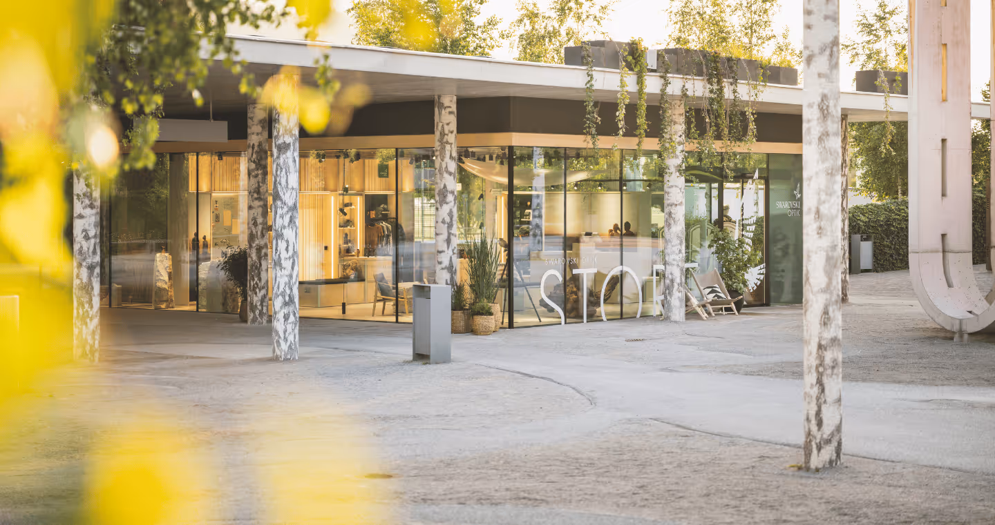 Exterior view of the Swarovski Optik Store at Swarovski Kristallwelten — modern glass building with open shop windows and natural design between birch trunks.