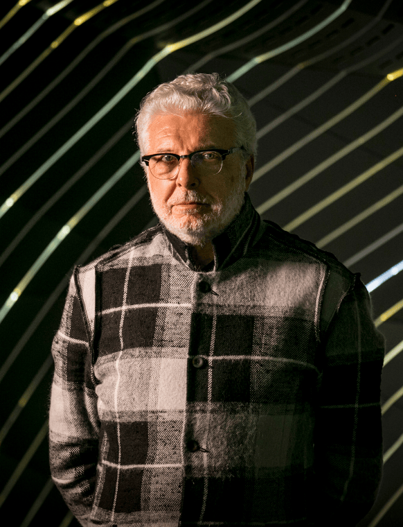 Porträt von André Heller mit weißem Haar, Brille und kariertem Jackett vor einem Hintergrund aus geschwungenen Lichtlinien.