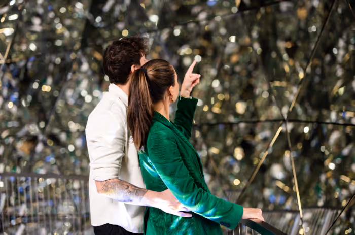 Paar im Kristalldom der Swarovski Kristallwelten: 595 Spiegeln funkeln im Licht, während die Frau nach oben zeigt und die Installation bestaunt.